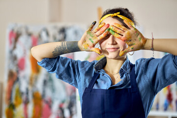 Portrait of creative woman holding her hands in paint over her eyes while creating abstract painting at home studio workshop