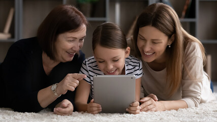 Wall Mural - Happy three female generations of family, girl, mom, grandma relaxing at home together, lying on heating comfortable floor, using tablet, watching online video on device, making call