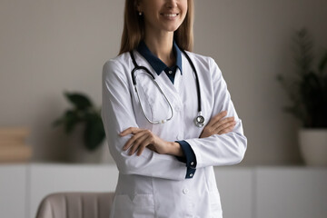 Happy smiling female doctor, hospital worker, physician, nurse wearing white coat standing in clinic office with folded hands. General practitioner, medical expert cropped portrait. Close up