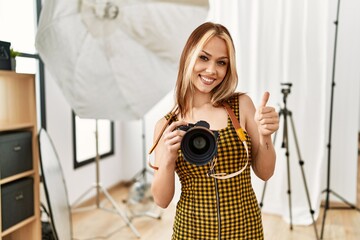 Young caucasian photographer girl holding professional camera at photography studio smiling happy and positive, thumb up doing excellent and approval sign