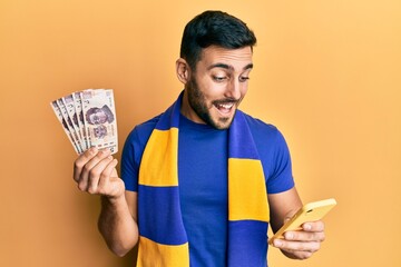 Poster - Young hispanic man football supporter using smartphone holding mexican pesos banknotes smiling and laughing hard out loud because funny crazy joke.