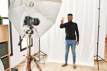 Wall Mural - Young hispanic man with beard posing as model at photography studio smiling looking to the camera showing fingers doing victory sign. number two.