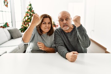 Wall Mural - Middle age hispanic couple sitting on the table by christmas tree angry and mad raising fist frustrated and furious while shouting with anger. rage and aggressive concept.