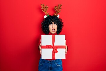 Sticker - Young middle east woman wearing deer christmas hat holding gift angry and mad screaming frustrated and furious, shouting with anger. rage and aggressive concept.