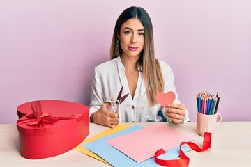 Young hispanic woman making valentine gift sitting on the table relaxed with serious expression on face. simple and natural looking at the camera.