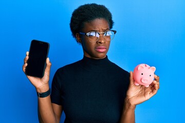 Canvas Print - Young african american girl holding piggy bank and smartphone clueless and confused expression. doubt concept.