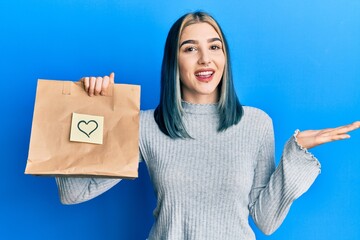 Wall Mural - Young modern girl holding take away paper bag with heart note celebrating achievement with happy smile and winner expression with raised hand