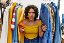 Young Hispanic Woman Searching Clothes On Clothing Rack Using Smartphone In Shock Face, Looking Skeptical And Sarcastic, Surprised With Open Mouth