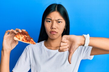 Poster - Young chinese woman eating tasty pepperoni pizza with angry face, negative sign showing dislike with thumbs down, rejection concept