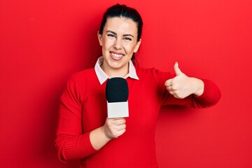 Poster - Young hispanic woman holding reporter microphone smiling happy and positive, thumb up doing excellent and approval sign