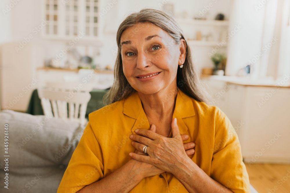 Middle-aged smiling woman feeling and showing appreciation of kindness. Elderly happy woman sitting in living room and looking at camera with with thanks. Thankfulness and gratitude concept - obrazy, fototapety, plakaty 