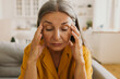 © shurkin_son - Middle-aged sad female sitting on sofa and looking down. Mature upset woman touching head and feeling pain in temples. Hard day and headache concept. Aging and health problems idea