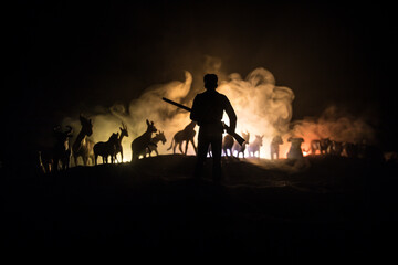 silhouette of a man (hunter) with rifle standing against group of animals in colorful dark backlight