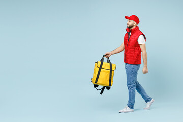 Wall Mural - Full size body length delivery guy employee man in red cap white T-shirt uniform work as courier hold yellow thermal food bag backpack isolated on pastel blue color background studio. Service concept.