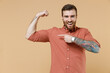 Strong sporty fitness assured tatooed young brunet man 20s with earrings wear apricot shirt show point on biceps muscles on hand demonstrate power isolated on pastel orange background studio portrait