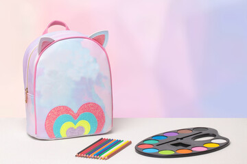 Back to school. A colourful school backpack, pencils or wooden crayons arranged in a row and watercolor paints on a white table over abstract bright background. Concept education. Space for your text 