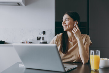 Young smiling housewife woman 20s in tshirt air pods eat breakfast talk video call use laptop pc computer work online listen to music cook food in light kitchen at home Healthy diet lifestyle concept