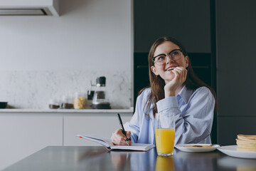 Wall Mural - Smart pensive wistful student young housewife woman 20s in blue shirt casual clothes glasses writing down recipe in notebook diary cook food in light kitchen at home Healthy diet lifestyle concept