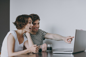 Young excited fun couple two woman man in casual t-shirt clothes sit table drink coffee point finger on laptop pc computer watch live stream in light kitchen at home together People lifestyle concept.