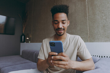 Wall Mural - Smiling happy confident young african american man in beige t-shirt siting t on grey sofa indoors apartment use mobile cell phone chatting online speak with friends rest on weekends staying at home.