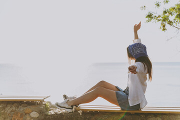 Side view full body young fun happy traveler tourist woman 20s in blue bandana shirt summer casual clothes rest sitting on wooden bench outdoors at sea beach People vacation lifestyle journey concept.