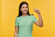 Smiling cute brunette young woman in mint tshirt standing and showing small size by fingers isolated over yellow background