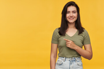 Wall Mural - Cheerful pretty young woman with dark hair in casual clothes pointing away to the side at empty space and looking at camera over yellow background