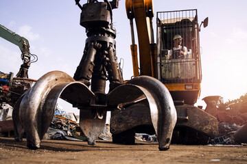 Man operating excavator industrial machine with claw attachment used for lifting scrap metal in junk yard.