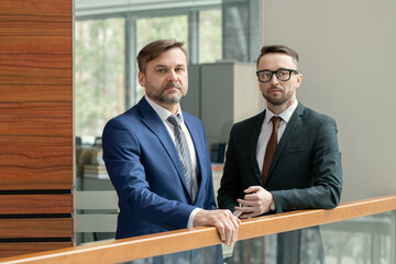 Poster - Portrait of two confident businessmen in elegant suits looking at camera while standing in the corridor of office