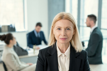 Sticker - Portrait of mature blonde businesswoman looking at camera while her colleagues sitting at meeting at office