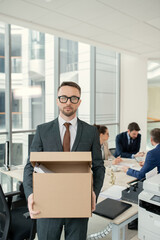 Aufkleber - Portrait of young businessman holding cardboard box with his stuff and looking at camera with business people sitting at meeting in the background