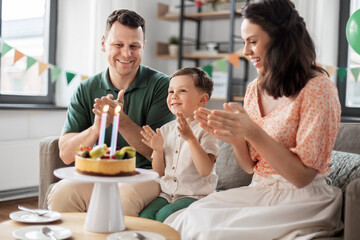 Wall Mural - family, holidays and people concept - portrait of happy mother, father and little son with four candles on birthday cake sitting on sofa at home party