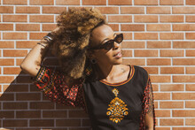 Beautiful African American Woman Wearing Sunglasses Leaning Against  Orange Bricks Wall Tiding Up Her Curly Hair. Copy Space For Text.