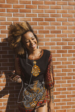 Beautiful African American Woman Leaning Against Orange Bricks Wall Wearing Earphones Listening To Music On Smartphone And Smiling. Copy Space For Text.