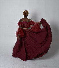 Wall Mural - Full length portrait of pretty African woman wearing long red renaissance medieval fantasy gown, standing pose on a light grey studio background.