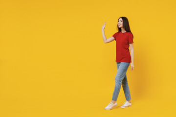 Wall Mural - Full size body length side view profile young brunette woman 20s wear basic red t-shirt stand go move step meet greet waving hand isolated on yellow background studio portrait. People emotion concept