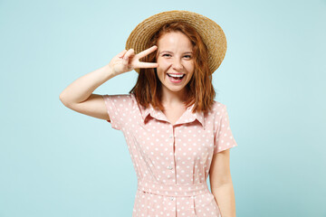 Laughing happy young redhead curly woman 20s show victory sign wears casual pink dress straw hat look camera isolated on pastel blue color background studio portrait. People emotions lifestyle concept