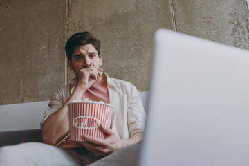 Poster - Young shocked confused man 20s wear casual clothes use laptop pc computer watch series hold takeaway popcorn bucket eat snack sit on grey sofa rest indoors at home on weekends People leisure concept