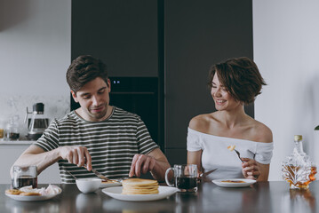 Wall Mural - Young cheerful fun happy couple two woman man in casual t-shirt clothes sit by table eat pancakes maple syrup have breakfast cook food in light kitchen at home together Healthy diet lifestyle concept.