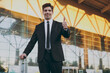 © ViDi Studio - Bottom view young traveler brunet businessman man in black classic suit stand outside at international airport terminal with suitcase valise show thumb up. Air flight business trip lifestyle concept.