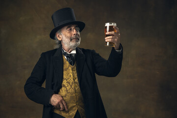 Elderly gray-haired man, gentleman, aristocrat or actor drinking beer isolated on dark vintage background. Retro style, comparison of eras concept.