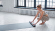 Slim blonde barefoot sportswoman in light beige tracksuit rolls rubber mat after training near large window