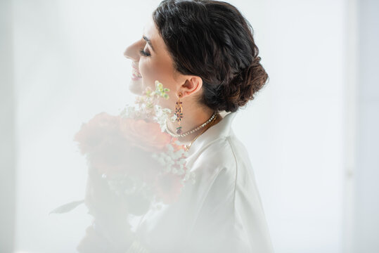 side view of happy indian bride with mehndi holding bouquet of flowers on white
