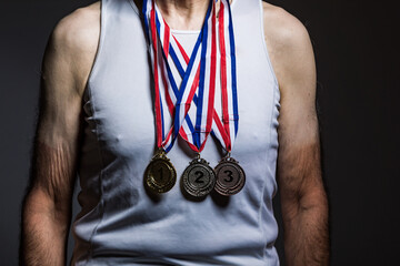 Elderly athlete wearing white tank top, with sun marks on the arms, with three medals on the neck, showing them, on a dark background. Sports and victory concept