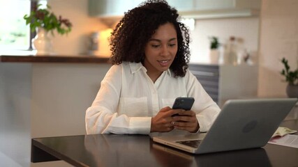 Poster - Joyful young mixed race woman chatting use smartphone enjoying break during work in front of laptop. Smiling female typing message or texting on mobile sitting on desk at home office