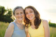 © Olga - Senior mother with gray hair with her adult daughter looking at the camera in the garden and hugging each other during sunny day outdoors, mothers day