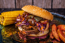 BRISKET BURGER SERVED WITH BREAD, BARBECUE, ONION, CHILI, PURPLE CABBAGE, POTATOES AND ELOTE ON A GREEN GLASS PLATE WITH WOODEN BACKGROUND
