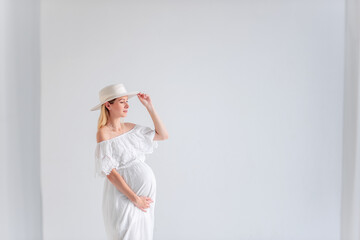 Wall Mural - Young pregnant woman in anticipation of a baby in a white hat, dress, hugs her belly. Tender blonde girl in a minimalistic, isolated interior of airy fabrics. Happy maternity pregnancy, surrogacy