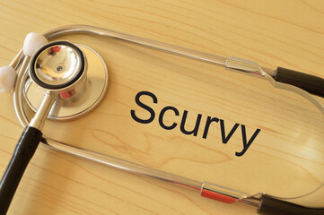 Stethoscope on the table with text SCURVY. Scurvy is a deficiency disease that results from lack of Vitamin C.