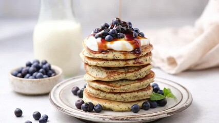 Wall Mural - Pouring maple syrup on stack of healthy poppy seed pancakes served with greek yogurt and wild blueberries